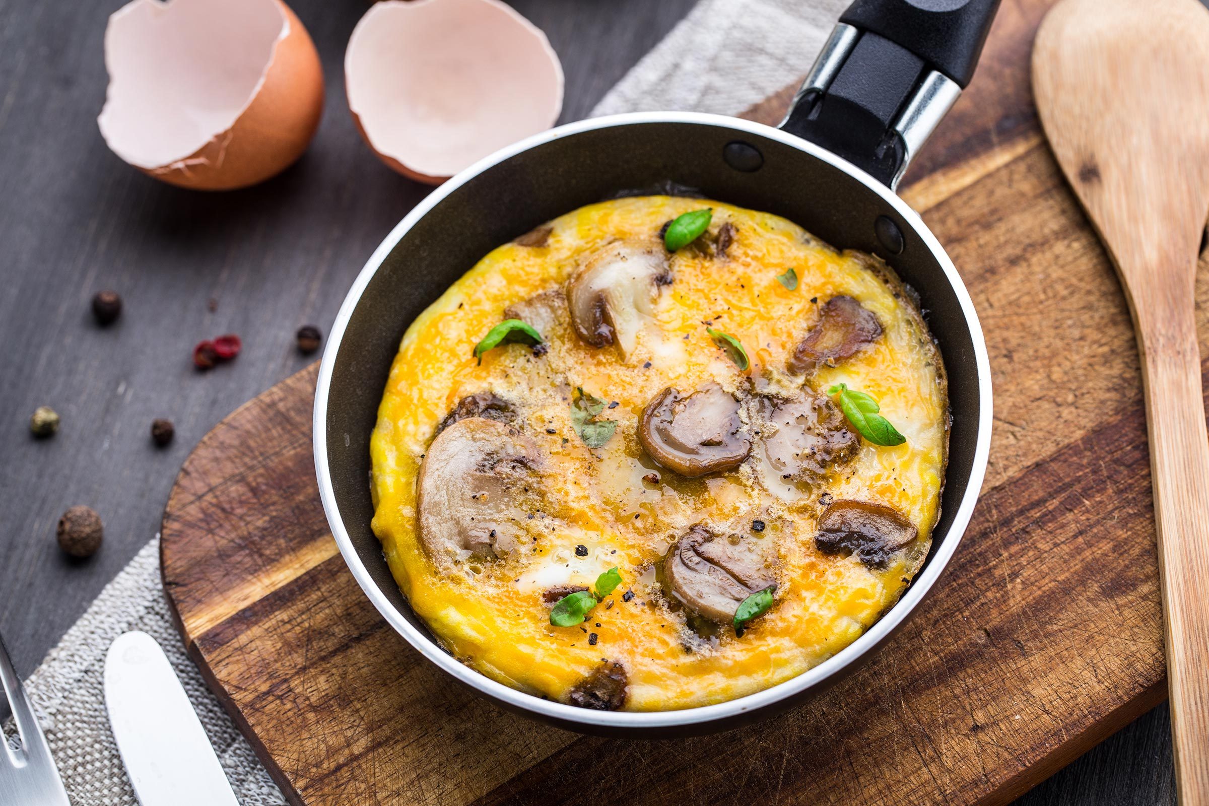 Omelette cooks in a small frying pan, surrounded by a wooden board, cracked egg shells, and a wooden spoon on a dark surface.