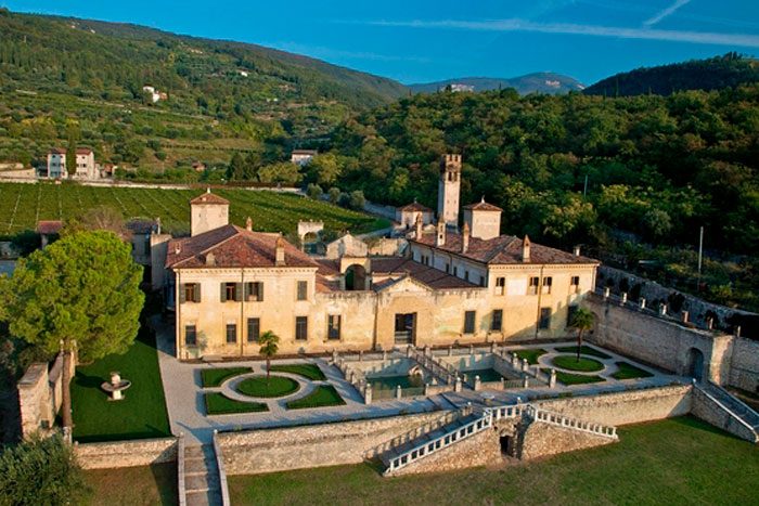 A historic mansion stands with a formal garden and fountains, surrounded by lush green hills and a distant mountain backdrop.