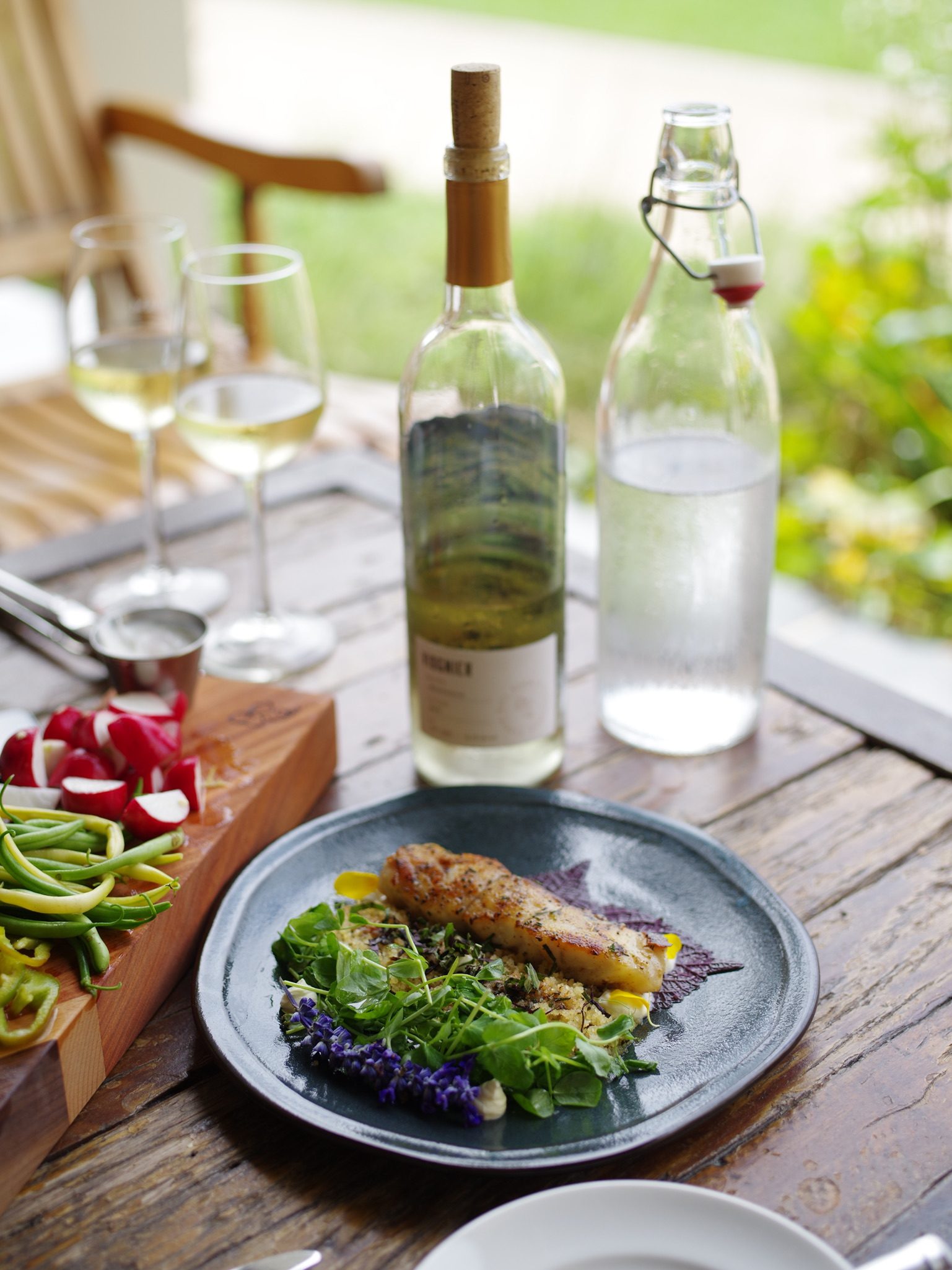 A plated meal featuring fish rests on a rustic table beside fresh vegetables, wine, and water bottles, set against a blurred outdoor backdrop.