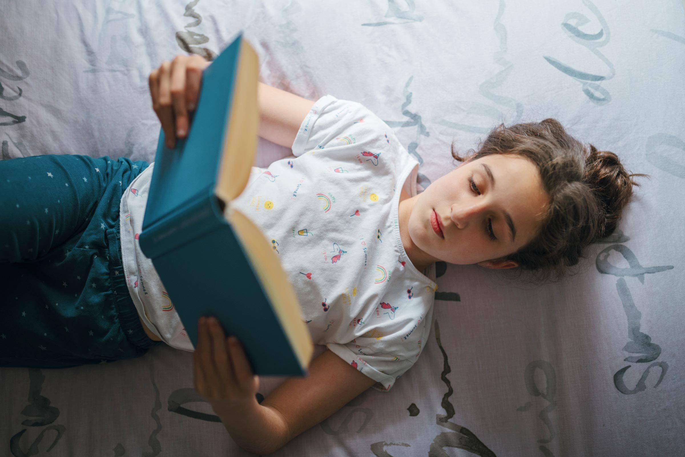 Person reads a blue book while lying on a bed, wearing a white shirt with colorful patterns.