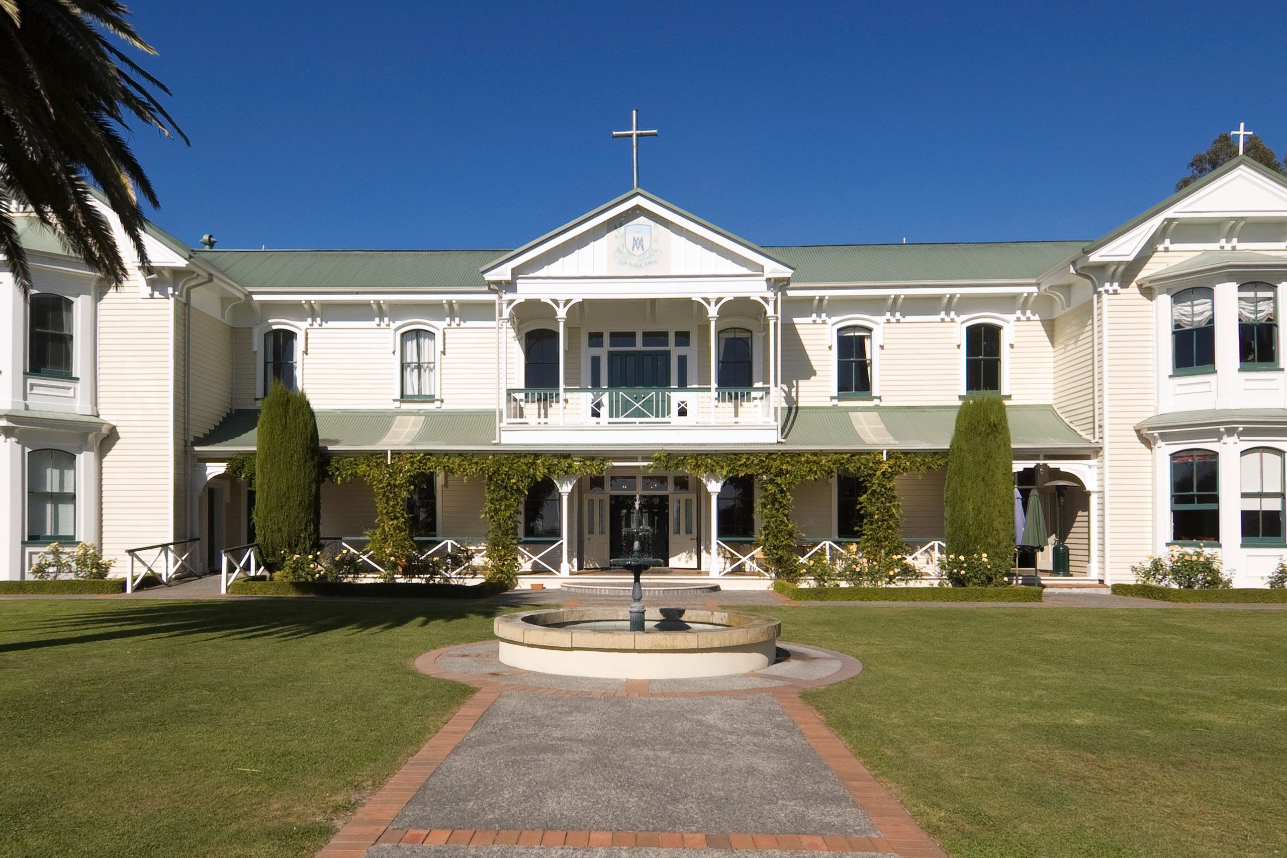 A white, two-story building with a cross stands, surrounded by green vines, manicured lawn, and a central stone fountain under a clear blue sky.