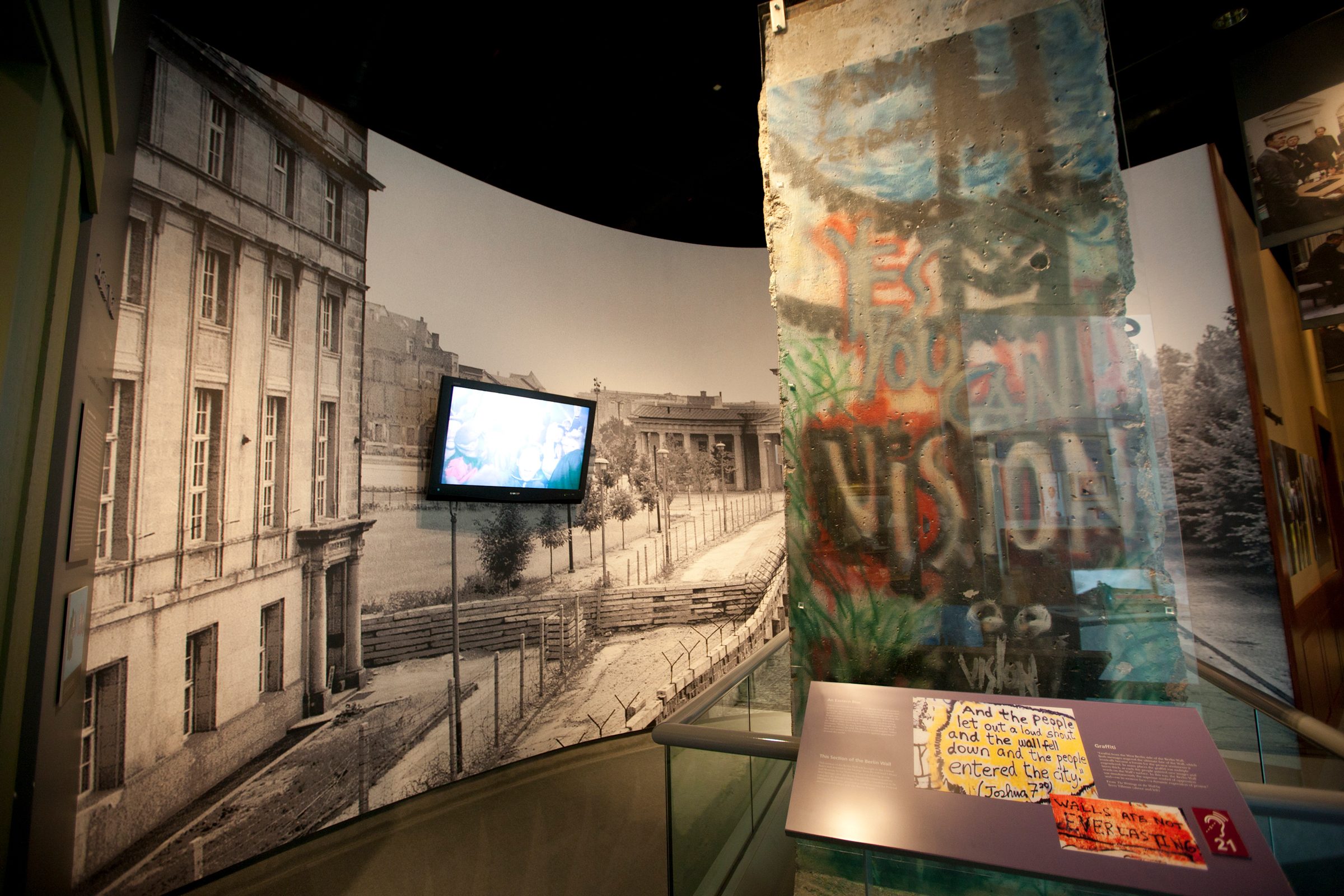Concrete wall fragment displays colorful graffiti: "Yes, you can vision." Mounted TV shows historical footage. Nearby, informational text on a panel set in a museum exhibit.