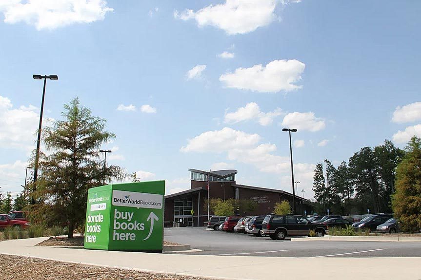 Sign promotes donating and buying books, standing beside a parking lot with several cars in front of a modern building and trees.
