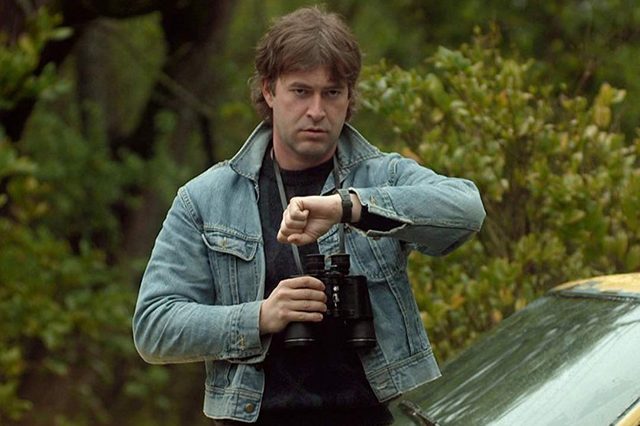 A person holds binoculars, checking their watch, standing beside a vehicle in a lush, green outdoor setting.
