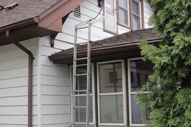 Ladder leans against a two-story house next to a tree, reaching the roof near a second-floor window.