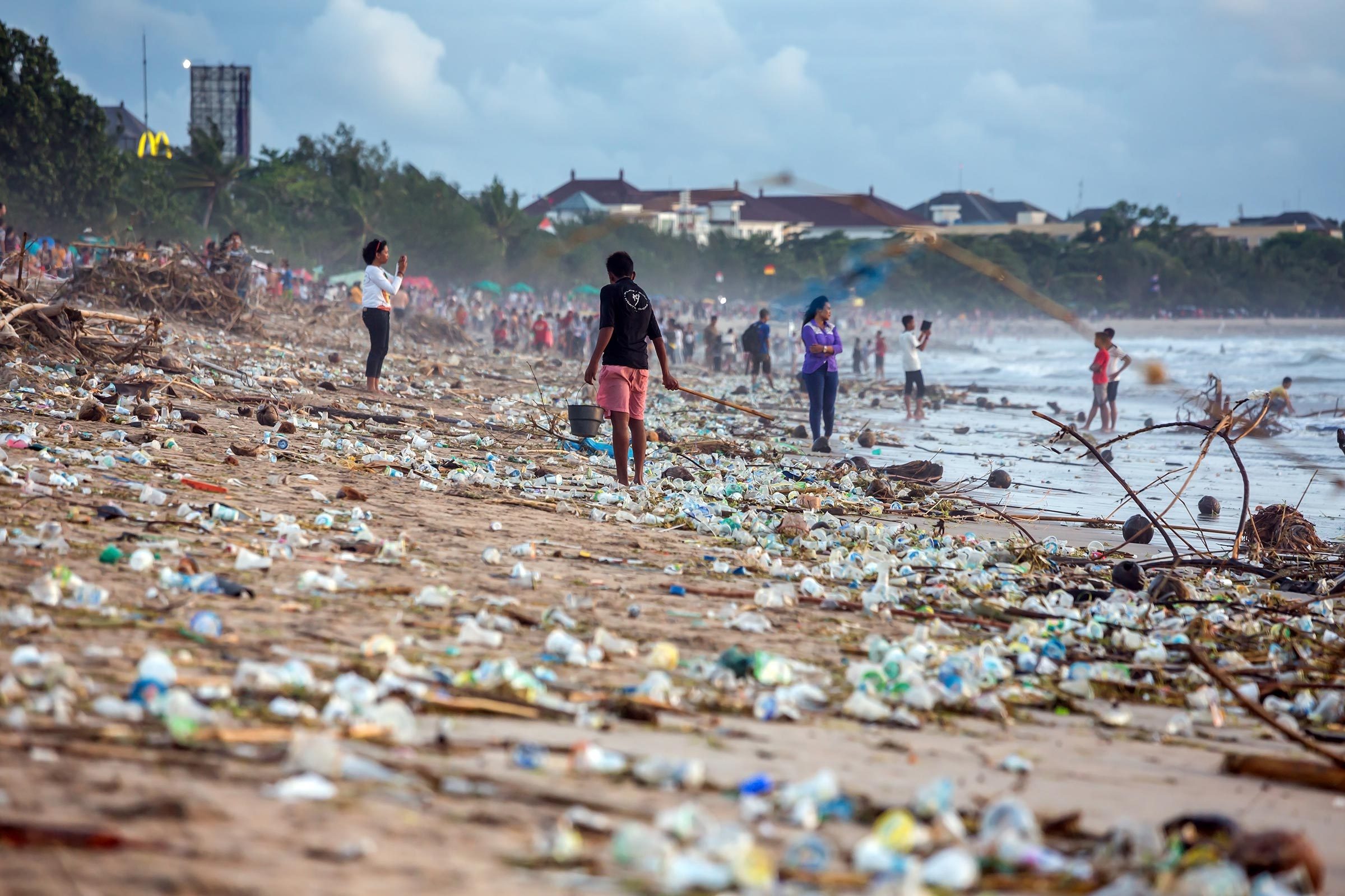 People walk on a littered beach, collecting trash with sticks, amidst scattered plastic and driftwood, under a cloudy sky.