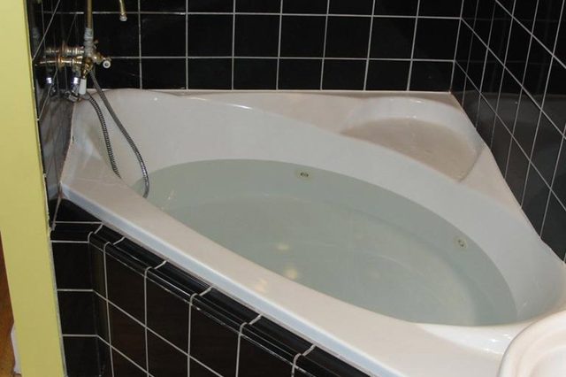Bathtub holds water, positioned in a corner, surrounded by black square tiles, with a gold faucet and wooden floor nearby.