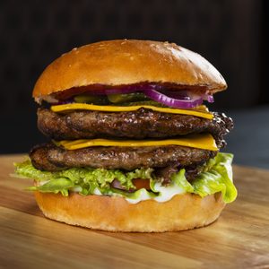 Burger stacked with two patties, cheese slices, lettuce, onions, and pickles sits on a wooden surface, against a dark background.