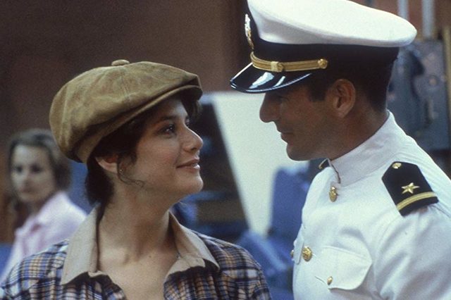 Woman smiles, wearing cap, looking at a man in a navy uniform; blurred background with another person, suggesting an indoor setting.