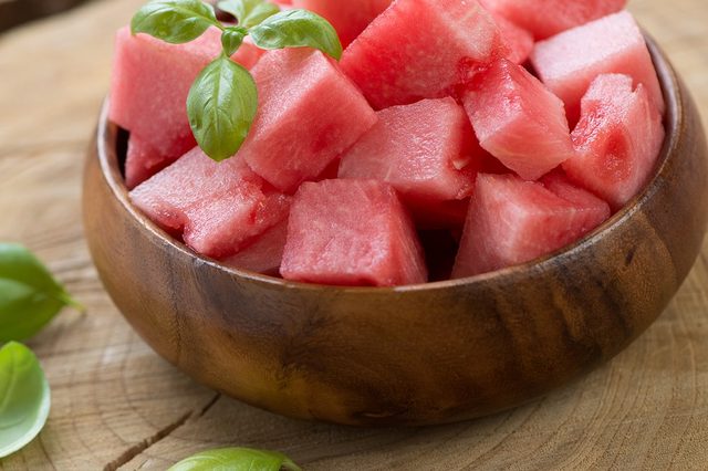 Wooden bowl holds watermelon cubes, topped with basil leaves, set on rustic wooden surface scattered with additional basil leaves.