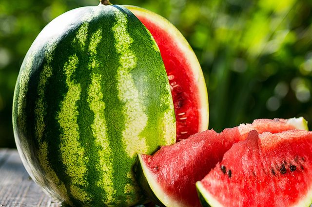 Watermelon sits sliced on a wooden table, revealing vibrant red flesh and seeds, with a lush green garden in the background.