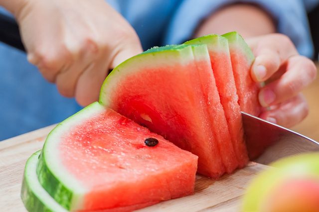 Hands slicing watermelon on a wooden cutting board, with other fruit blurred in the foreground.