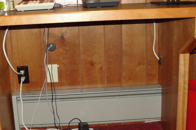 Power strip lies on red carpet, plugged into wall, beneath a wooden desk holding a calculator and a phone in a paneled room.