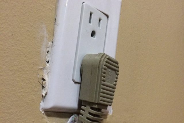 Plug loosely inserted into a damaged electrical outlet, with frayed cable, on a beige wall.