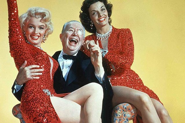 Three people pose joyfully, two women in red sequined dresses and a man in a tuxedo, against a yellow background.