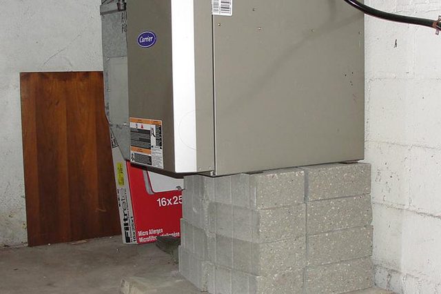 Furnace rests on stacked concrete blocks in a basement, adjacent to a white brick wall and a wooden board.