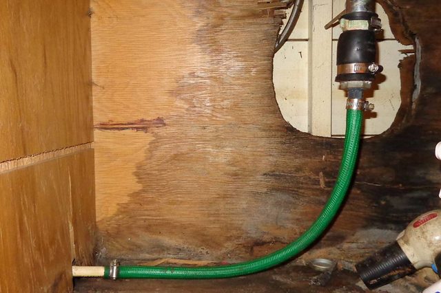 Green hose connected to plumbing under a sink, surrounded by worn, discolored wood and various tools in a cabinet.