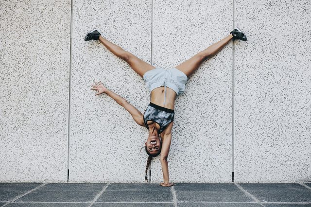 Person doing a one-handed handstand against a speckled wall, legs spread apart, wearing athletic clothing, smiling widely, on a tiled surface.