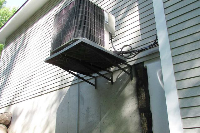 Air conditioning unit mounted on wall, cables attached, outside house with siding; sunlit and surrounded by greenery and mulch.