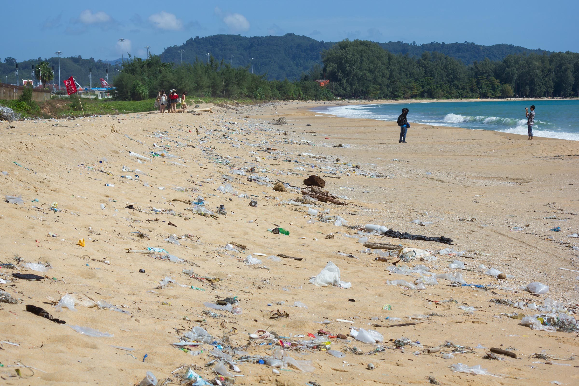 Trash scatters across a sandy beach; people walk and take photos in the background, surrounded by forested hills and ocean waves.