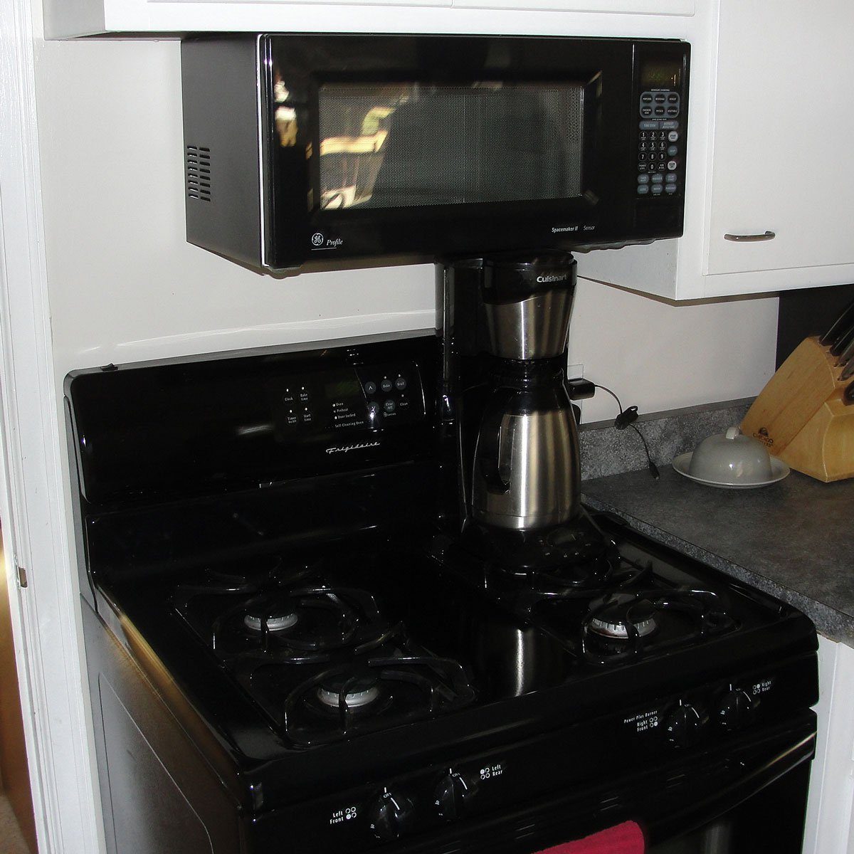 Coffee maker sits on a stove beneath a microwave, in a kitchen with countertops and cabinets.