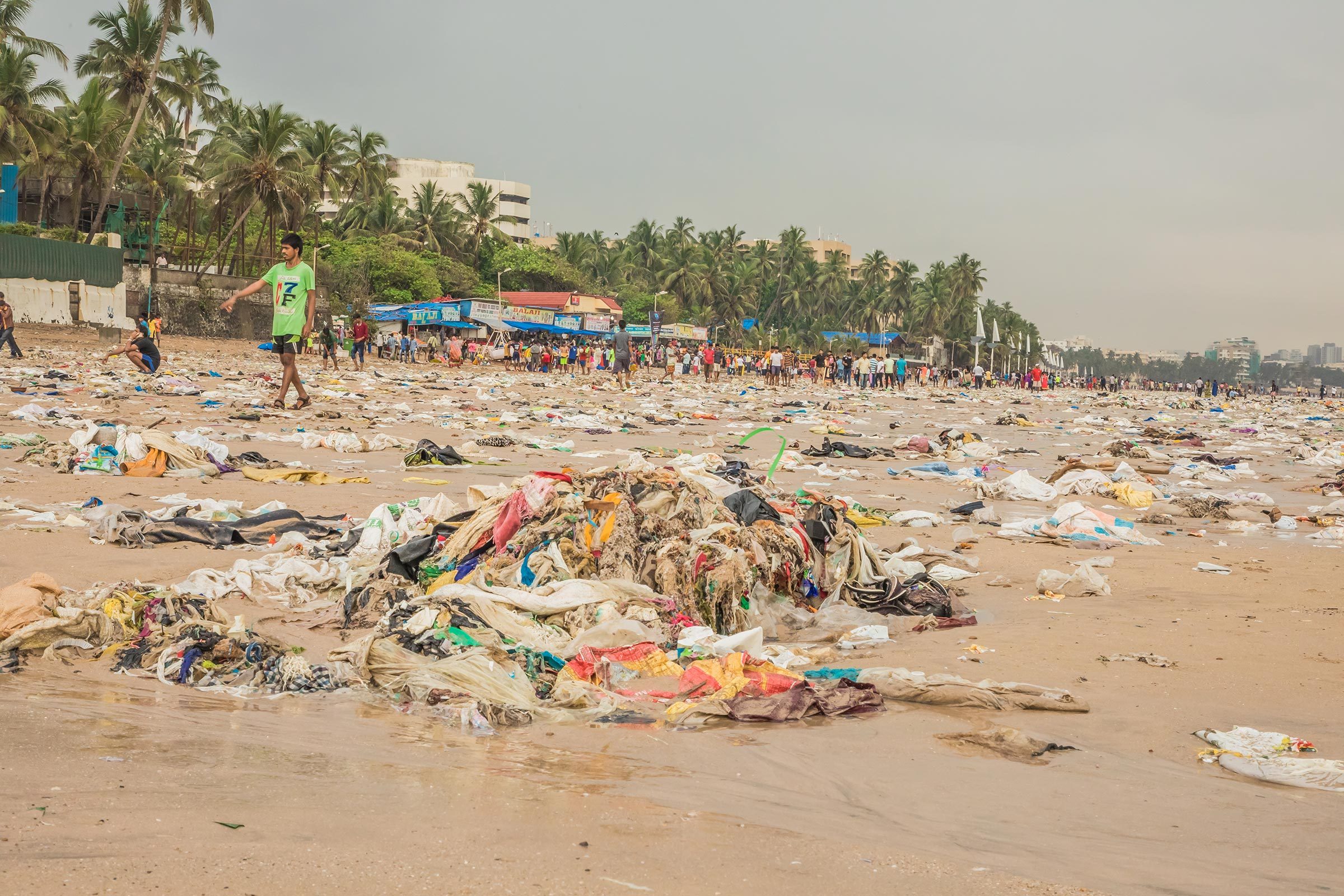 Trash litters the sandy beach shoreline, where people walk and palm trees line the background, creating a crowded and polluted seaside environment.