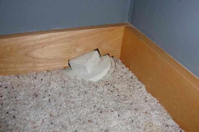 Outlet, partially covered by carpet, located in a corner with wooden baseboards and gray-painted walls.
