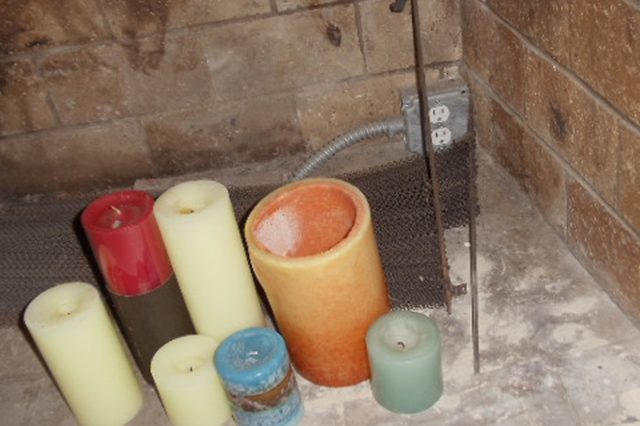 Candles are arranged on a fireplace hearth near a metal grate and electrical outlet, surrounded by brick walls.