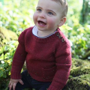 A young child smiles, sitting on mossy ground, surrounded by lush green foliage and sunlight filtering through trees.