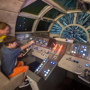 Children operate spaceship controls, simulating light-speed travel in a spacecraft cockpit with numerous buttons and glowing displays, surrounded by a starry space backdrop.