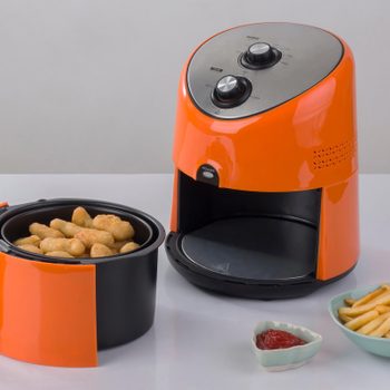 Air fryer cooks nuggets beside a plate of fries and ketchup on a white table.