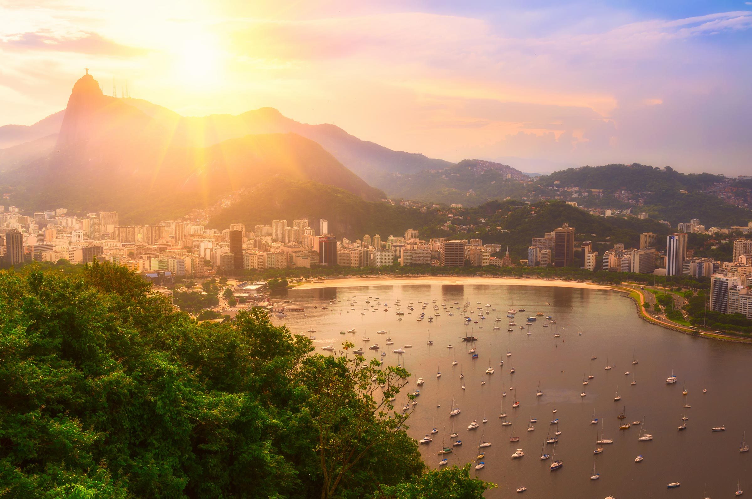 Mountaintop statue overlooks bay with many boats; foreground has lush greenery, background shows city at sunset, with vibrant sky and distant hills.