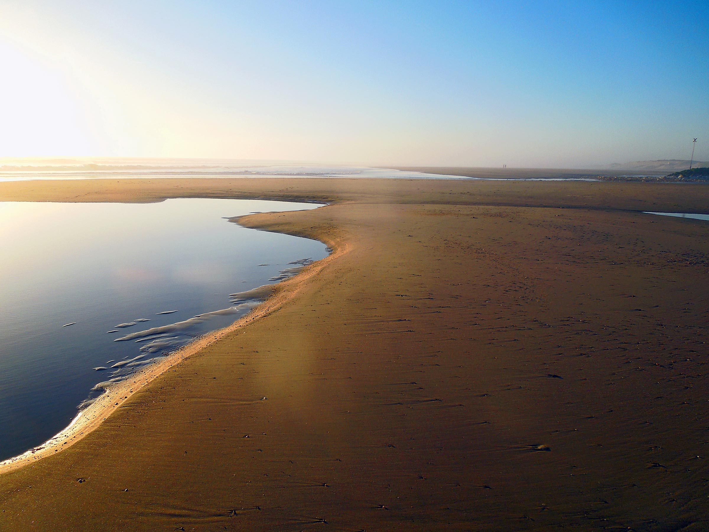 Sandbar curves along a tranquil blue tide pool under a clear sky at dawn, with distant horizon blending into the light.