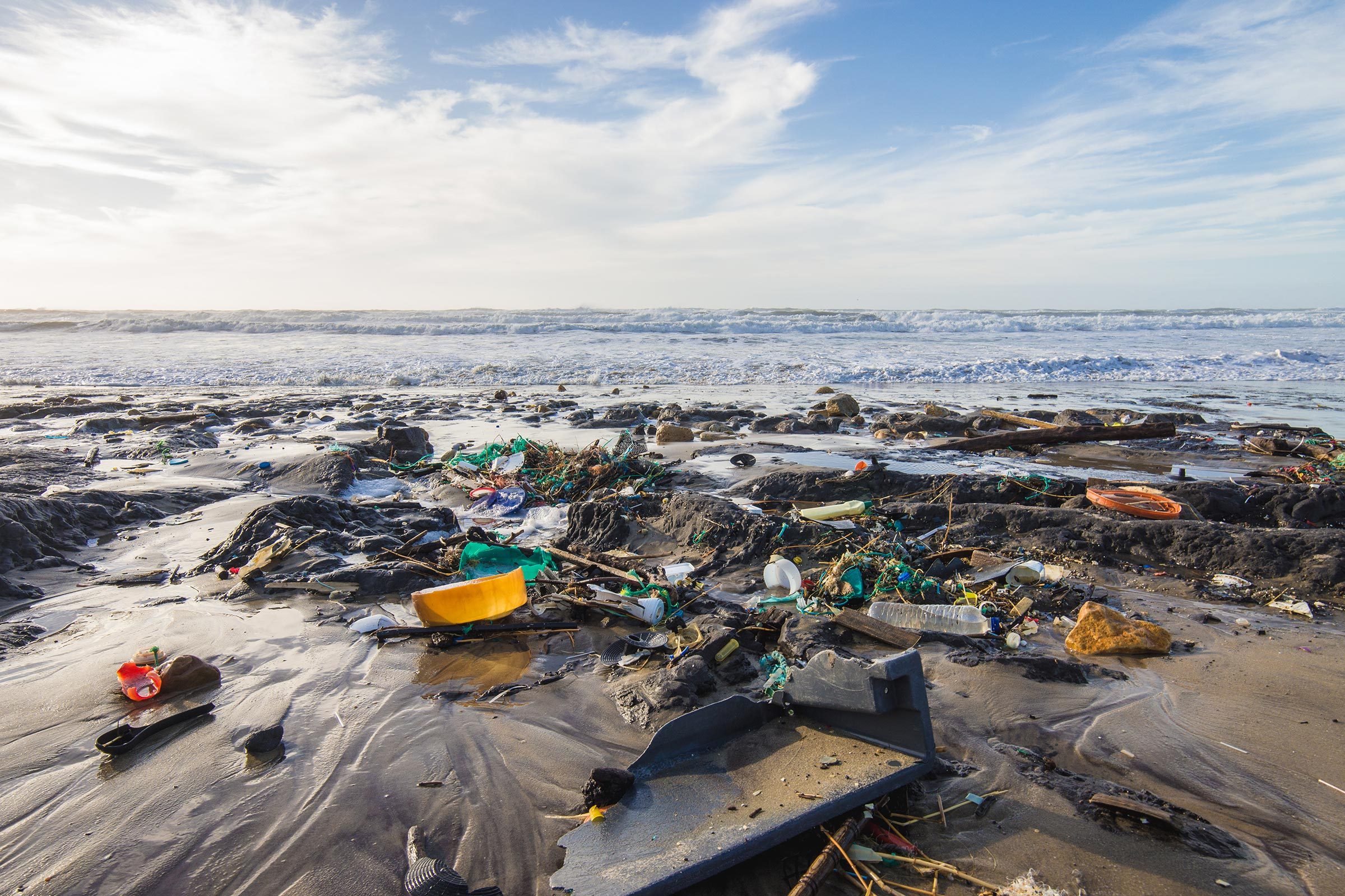 Debris litters the shore, scattered across wet sand and rocks, with ocean waves crashing under a cloudy sky.