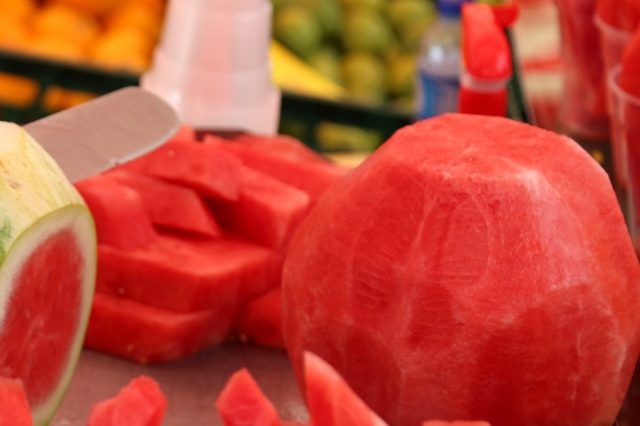 Whole and sliced watermelon displayed on a market stand, surrounded by yellow and green fruits in the background.