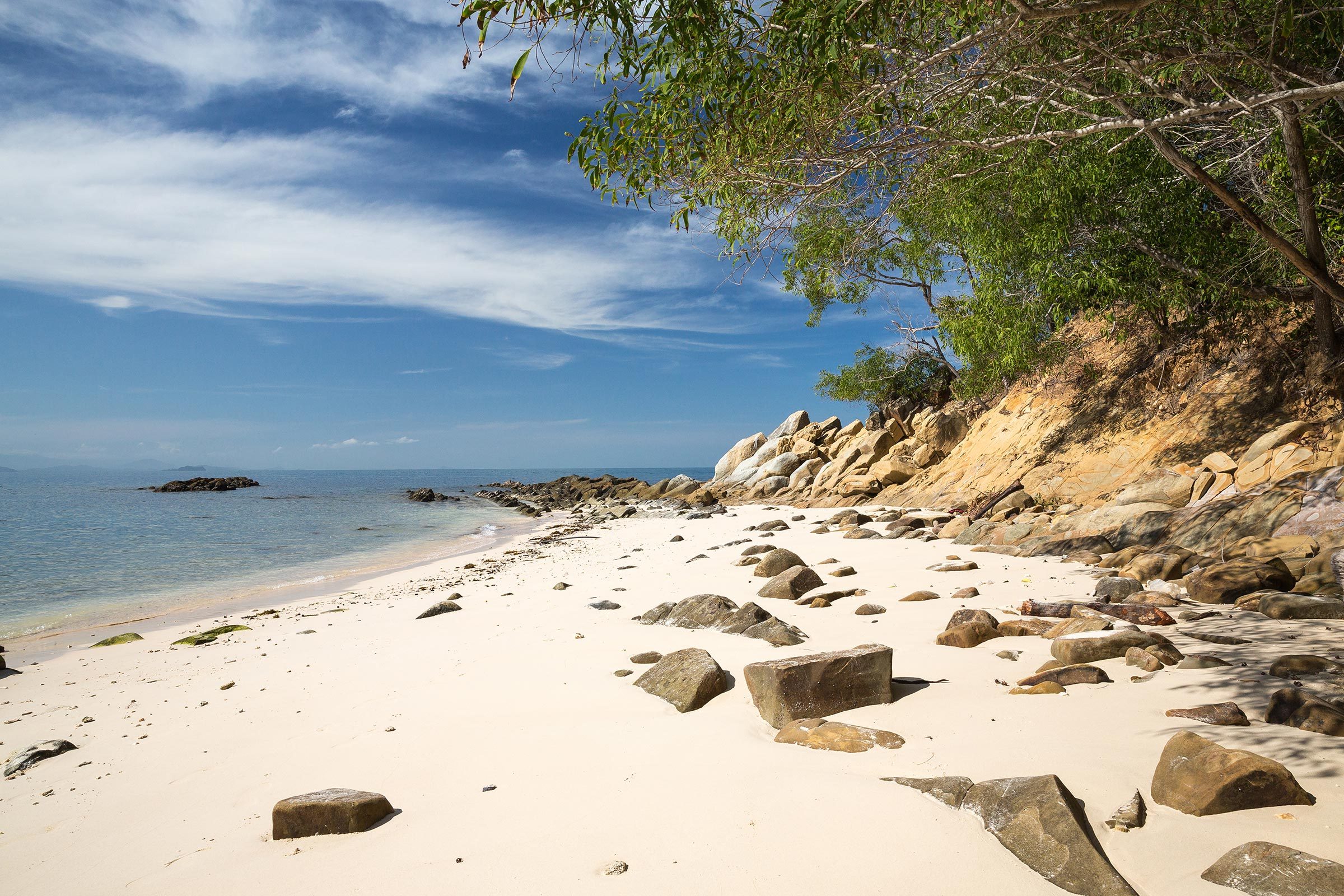 Beach, lined with scattered rocks, curves beside calm sea; trees overhang, casting shadows on sand under a clear, partly cloudy sky.