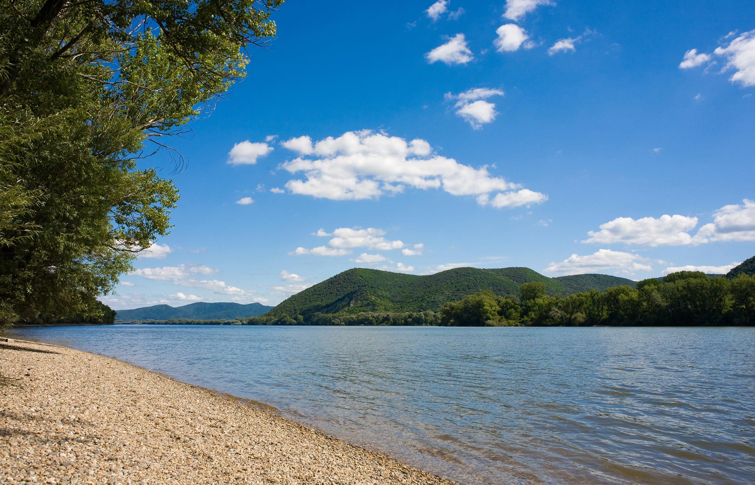 Pebble beach meets calm lake; mountains and trees surround under a bright, cloud-dotted blue sky.