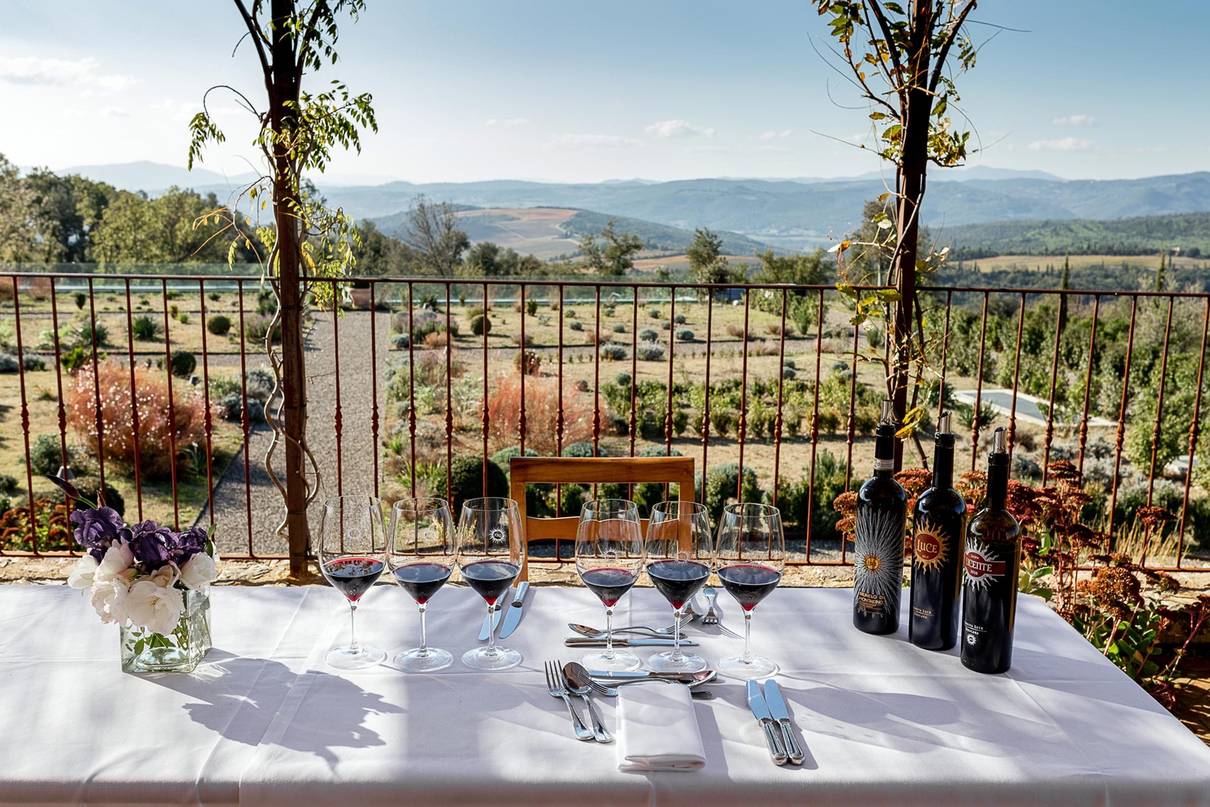 Wine glasses and bottles rest on a table with a white cloth overlooking a scenic vineyard and rolling hills under a clear blue sky.