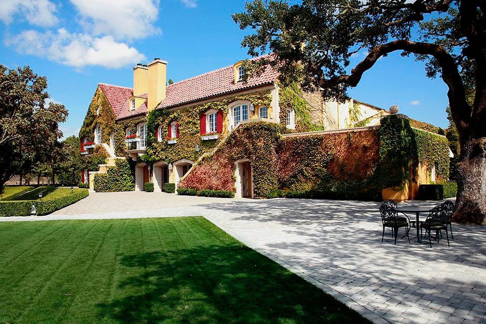 A villa with ivy-covered walls stands under a clear sky, surrounded by trees, and bordered by a stone patio and green lawn.