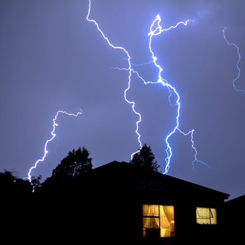 Lightning streaks across a dark sky, illuminating a silhouette of trees and a house with a lit window.