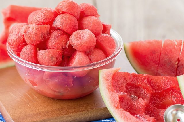 Watermelon balls fill a clear bowl, sitting on a wooden board with sliced watermelon nearby, atop a blue-striped cloth.
