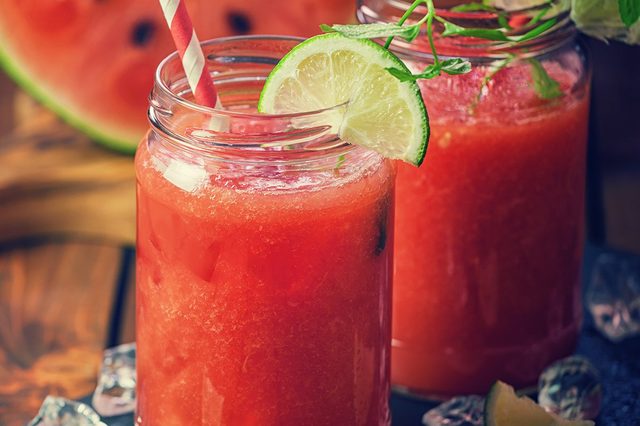 Two mason jars hold watermelon juice with lime slices, sitting amidst ice cubes on a dark surface, accompanied by a watermelon slice.