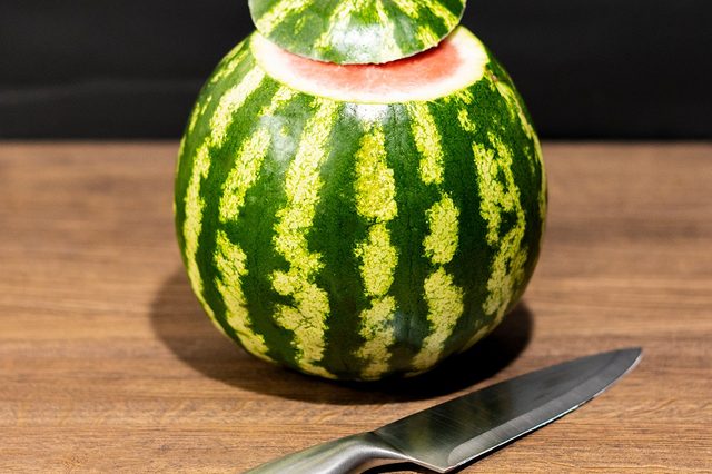 A watermelon with its top removed sits on a wooden table beside a shiny knife; dark background.
