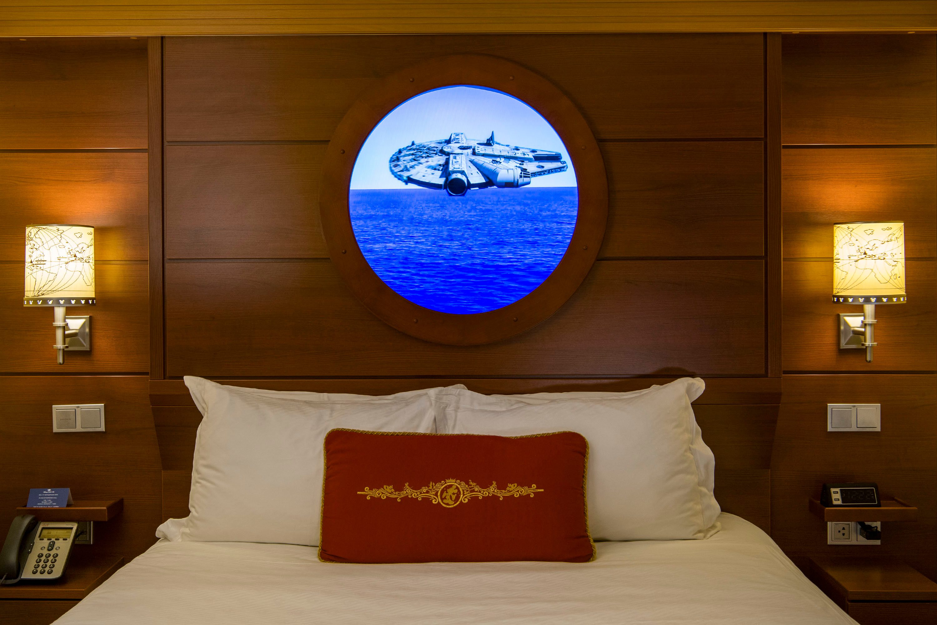 A spaceship is displayed on a digital round window above a neatly made bed, flanked by lit decorative lamps.