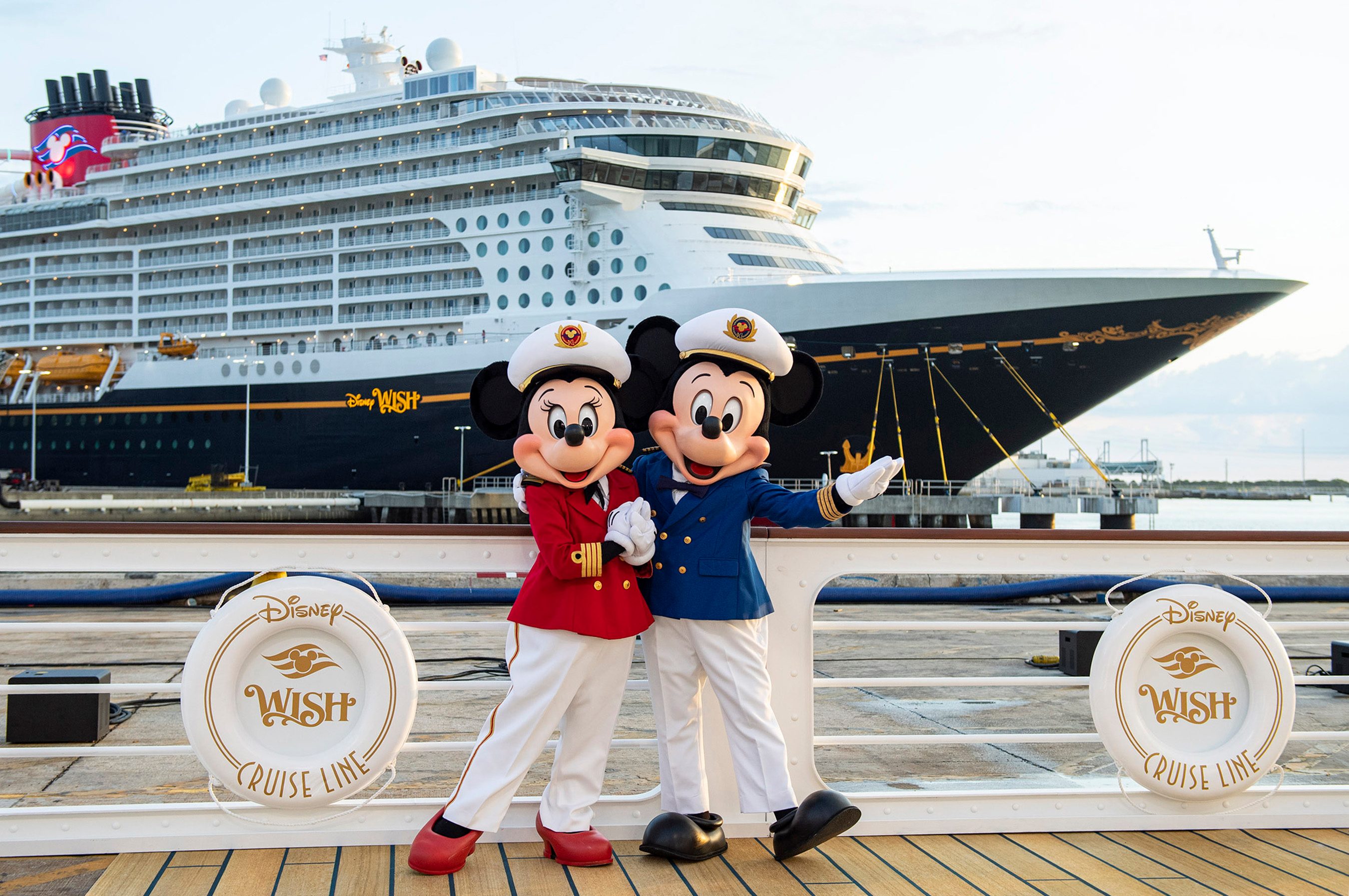 Characters in captain uniforms pose on dock, with a large cruise ship in the background. Lifesaver reads "Disney Wish Cruise Line."