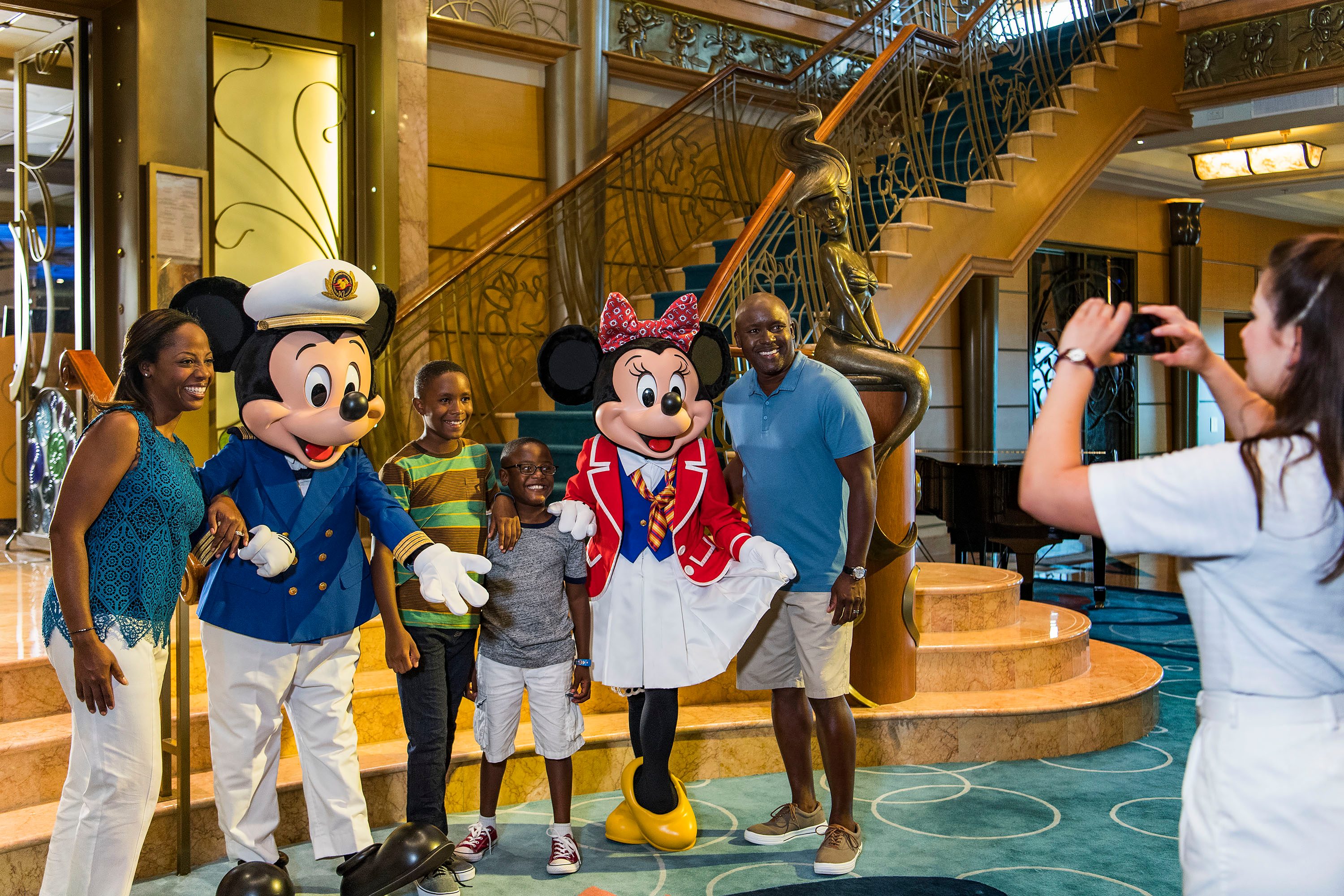 Characters and people pose for a photo, smiling in a grand interior with a decorative staircase and statue.