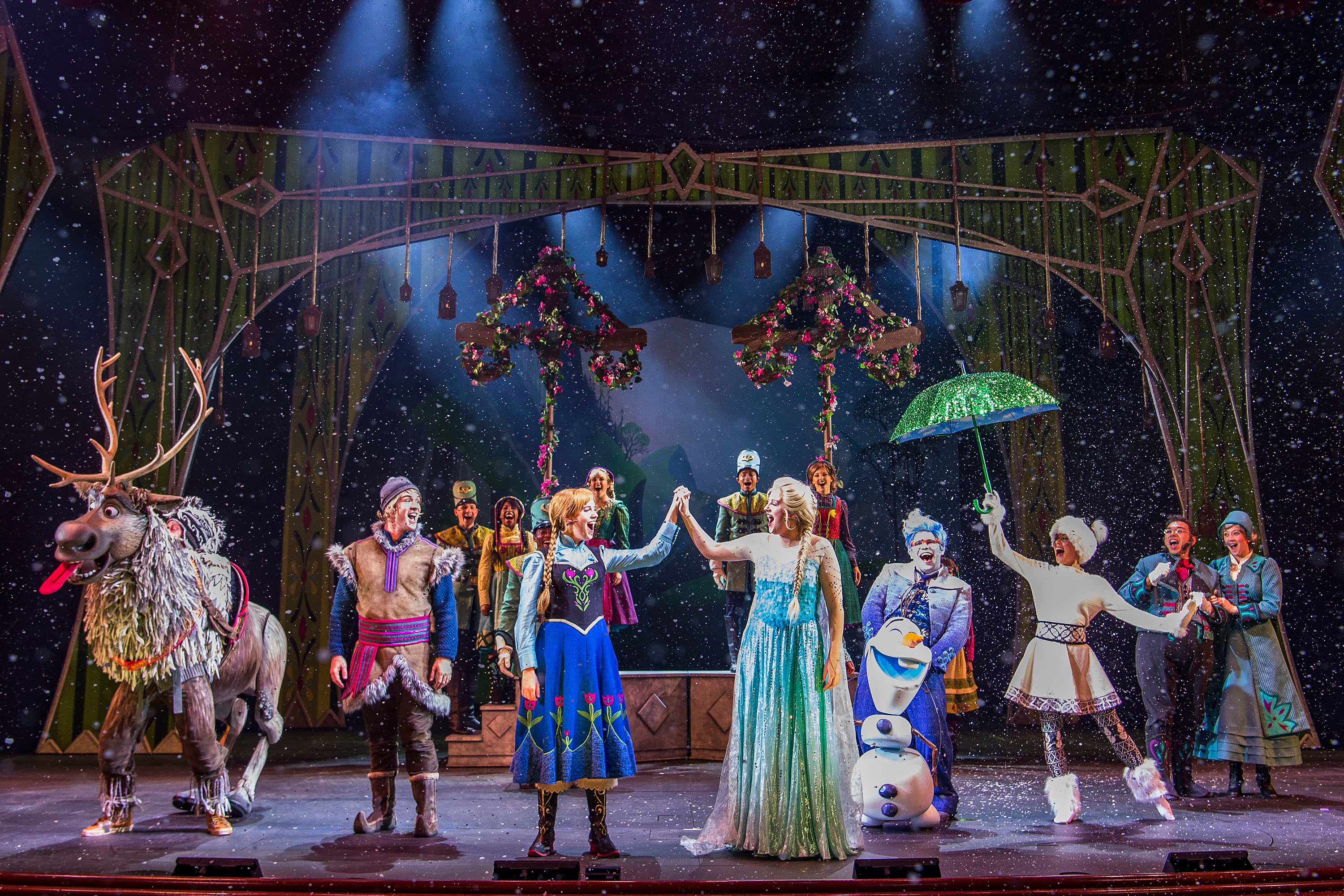 Characters perform joyfully in a winter-themed stage setting, with snow falling and colorful costumes, under dim theatrical lighting.