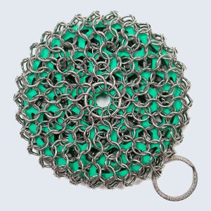 Metal chainmail scrubber rests on green sponge, circular shape, against a plain background. A metal ring at the edge reads 