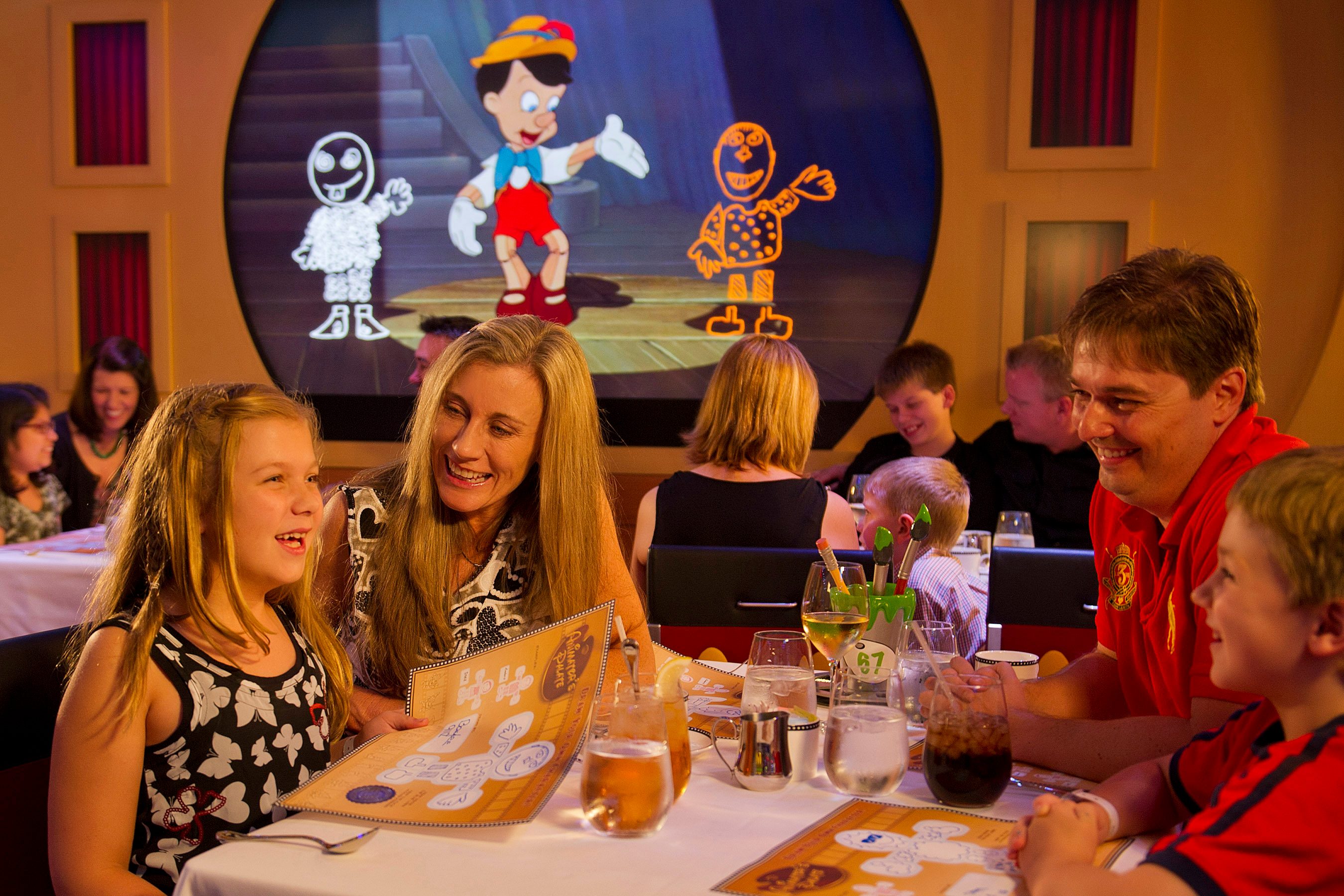 Family dining, smiling together while watching cartoon characters on a screen, in a lively restaurant setting.