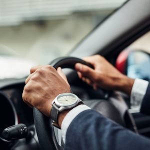 Hands grip a car steering wheel, wearing a watch, inside a vehicle. A blurred exterior is visible through the window.